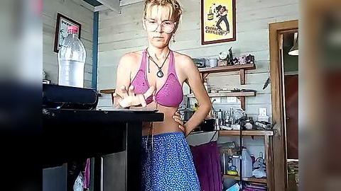 Media: Video of a young woman with glasses and short blonde hair, wearing a pink bikini top and blue patterned skirt, standing in a cluttered, dimly lit room with wooden walls and shelves.