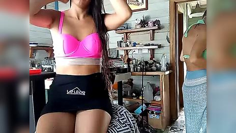 Media: Video of a young woman with long, dark hair in a vibrant pink sports bra and black high-waisted shorts, standing in a cluttered, dimly lit room with a white wooden shelf and various objects.