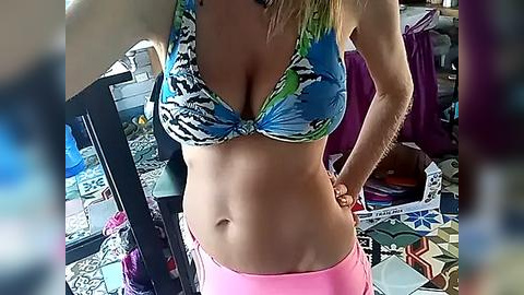 Media: Video of a slender woman with light skin, wearing a colorful zebra-print bikini top and pink shorts, standing in a cluttered room filled with scattered papers and a black desk.