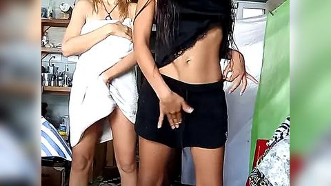 Media: Video of two women, one in a towel, the other in black shorts, in a casual, cluttered room with a green shower curtain and patterned bedding.