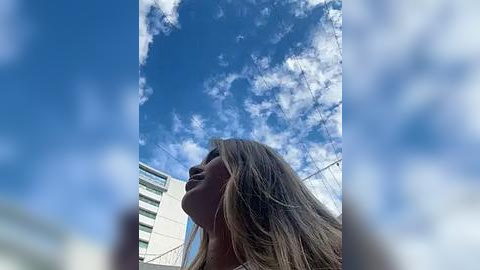 Media: A video of a blonde woman gazing upward, her long hair cascading over her shoulders, against a bright blue sky with scattered clouds.