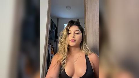 Media: Video of a light-skinned woman with long, wavy blonde hair, wearing a black tank top that accentuates her large breasts, posing in a bedroom doorway. Background shows a cluttered room with clothes hanging.