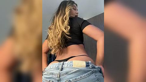 Media: Video of a curvy, light-skinned woman with long, wavy blonde hair, wearing a black crop top and denim shorts, exposing her black thong, viewed from behind.