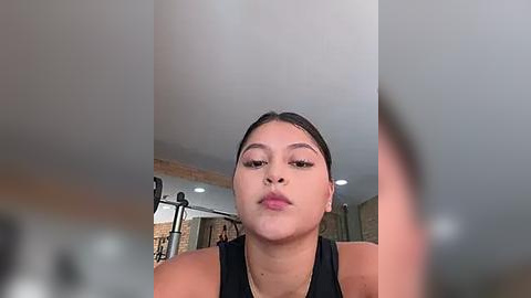 Media: Video of a young Latina woman with medium skin tone and dark hair, wearing a black tank top, puckering her lips for a selfie. Background features a gym with equipment and a brick wall.