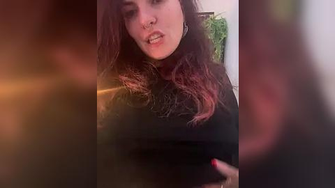 Media: Video of a young woman with long, dark hair, wearing a black turtleneck, and a nose ring, indoors with blurred background and warm lighting.