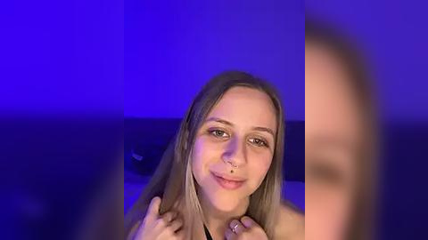 Media: Video of a smiling Caucasian woman with long blonde hair, wearing a nose ring, against a purple-lit background, holding her collar.