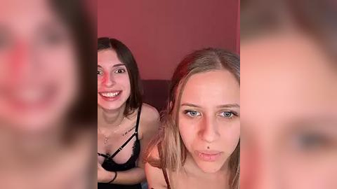 Media: Video of two smiling young women in lingerie, one with light skin, straight brown hair, and small breasts, the other with blonde hair, blue eyes, and medium breasts, set against a red background.