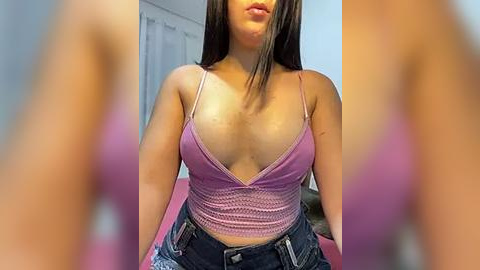 Media: Video of a young, light-skinned woman with long, straight brown hair, wearing a low-cut, purple crop top that reveals cleavage and high-waisted, ripped blue jeans. Background features a pink bed and white curtains.