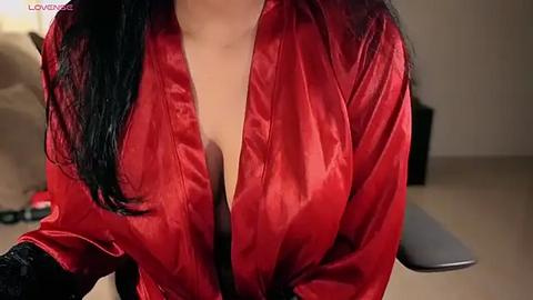 Media: Video of a woman with long black hair wearing a deep red satin robe, revealing cleavage. Background shows a beige sofa and a blurred red object.