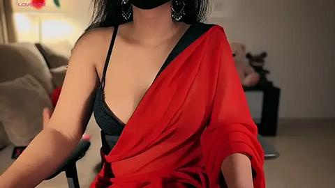 Media: Video of a woman in a red sari, black bra visible, dark hair, wearing a black mask, sitting in a living room with beige sofa and soft lighting.