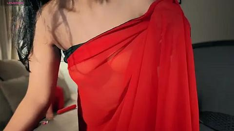 Media: Video of a woman with light brown skin, wearing a sheer red saree draped over her left shoulder, revealing a black bra strap, in a modern, dimly-lit room.