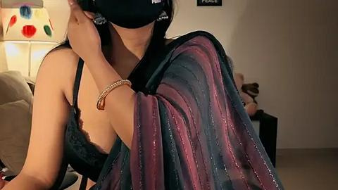 Media: Video of an Asian woman with medium skin tone, wearing a black mask and dark green lace bra, adjusting a purple sari in a dimly lit room with a beige sofa, colorful wall art, and a partially visible person in the background.