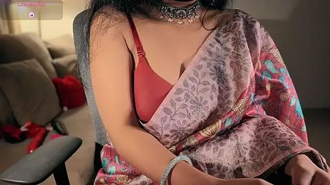 Media: Video of a South Asian woman in a red bra, draped in a floral-patterned pink sari, wearing a statement necklace. Indoor setting with beige couch and scattered clothes in the background.