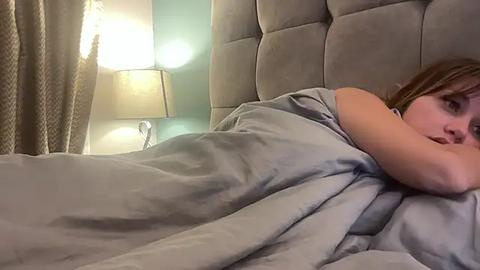 Media: Video of a young Caucasian woman with light skin and straight brown hair, lying in bed under a gray duvet, looking relaxed. The bed has a tufted headboard. A beige lamp with a white shade is on the nightstand.