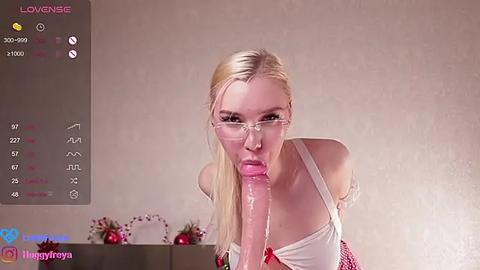 Media: Video of a blonde woman with glasses performing oral sex on a large, erect dildo. She wears a white strapless top. Background shows a simple room with a digital interface displaying \"Lovense\" and \"Meggymay.\