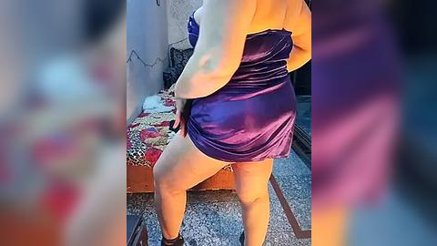 Media: Video of a curvy woman in a purple satin dress, standing with her back to the camera, highlighting her full buttocks and legs. Background includes a patterned couch, a cluttered room, and a soft-focus, blurry effect.