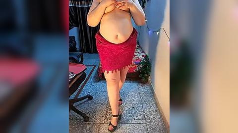 Media: Video of a plus-sized woman with fair skin, wearing a red lace skirt and black high heels, standing in a dimly lit indoor setting. She covers her breasts with her hands.