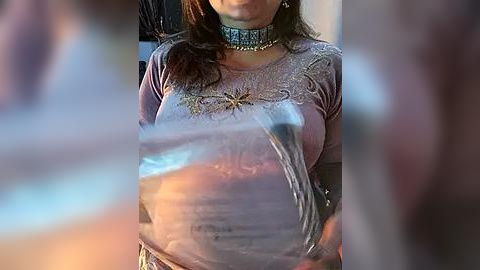 Media: Video of a woman with long brown hair, wearing a silver glittery top with a star design, and a choker necklace. She is holding a plastic bag filled with water, partially obscuring her face.