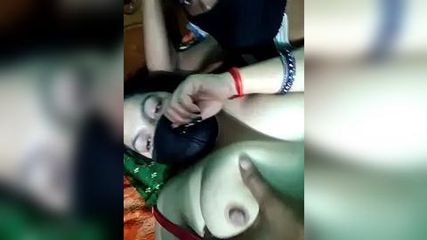 Media: Video of a woman with fair skin and medium breasts, wearing a red bracelet, being gagged with a black object, lying on a green and orange patterned surface.