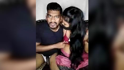 Media: Video of a shocked Indian man in a black t-shirt, sitting on a couch, watching a woman in a red dress unhook her bra.