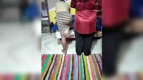 Media: Video of a woman in a black and white striped dress kneeling on a colorful, multi-striped rug, surrounded by other people in the background.