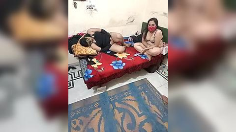 Media: A video of two women in a small, dimly-lit room with tiled floors, one wearing a red mask and a beige robe, the other lying on a red bedspread with blue floral patterns.