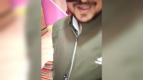 Media: Video of a smiling man with light skin, wearing a green jacket and sunglasses, standing in a room with a pink wall and striped rug.