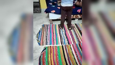 Media: Video of two individuals standing on a vibrant, multi-colored striped rug, in a brightly lit room with a blue sofa and colorful pillows in the background.