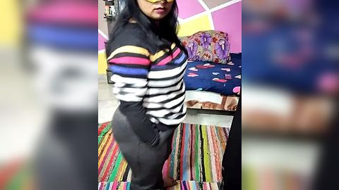 Media: Video of a young woman with long black hair, wearing a striped sweater and black pants, standing in a colorful, patterned room with a striped rug.