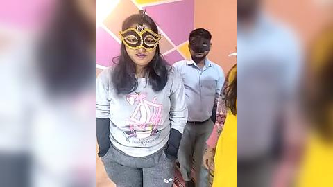 Media: Video of a woman in a light gray shirt with a graphic design, wearing yellow goggles and gray pants, walking through a pink and orange geometric patterned room.