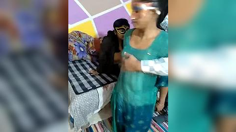 Media: Video of a young boy in a teal shirt, wearing a mask, standing near a bed with a black-and-white striped blanket in a room with multi-colored walls and a striped rug.