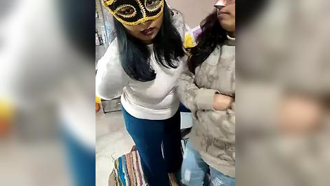 Media: Video of two women in a dimly lit room; one in a white sweater and blue pants, wearing a yellow mask, and the other in a beige jacket.