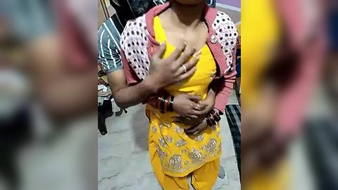 Media: Video of a young woman with medium skin tone, wearing a bright yellow sari with intricate embroidery, a pink and white patterned jacket, and a black bracelet, standing indoors.