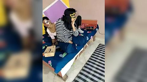 Media: A video of two children in a playful, colorful bedroom. One child wears a black-and-white striped shirt, black pants, and a yellow mask, sitting on a bed with a blue blanket. The other child, in a pink shirt, sits beside them on the bed.