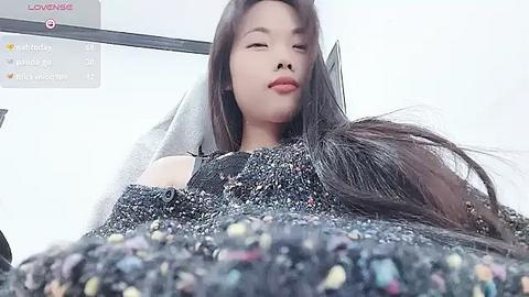 Media: Low-angle video of a young Asian woman with long, dark hair, wearing a black, textured sweater, in a brightly lit room, with a virtual background displaying \"Lovesense\" and \"Mia.\