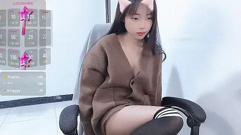 Media: A video of an East Asian woman with long black hair, wearing a brown cardigan and striped knee-high socks, sitting on a chair in a minimalist room.