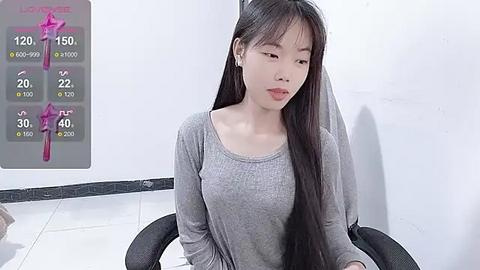 Media: Video of a young Asian woman with long black hair, wearing a grey long-sleeve top, seated indoors. Weather forecast screen shows rain and high temperature.