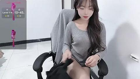 Media: A video of an East Asian woman with long black hair, wearing a grey long-sleeved top, sitting in a black office chair. She's pulling up her black skirt, revealing her legs. Background includes a pink vibrator on a glass desk.