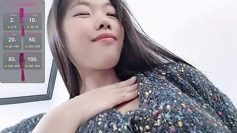 Media: Video of an East Asian woman with long black hair, fair skin, and a relaxed, closed-eye expression. She wears a multicolored knitted sweater. Background includes a white wall and a digital thermometer with a temperature reading of 100.9\u00b0F.