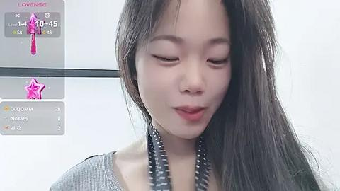 Media: Video of a young East Asian woman with long, straight black hair, wearing a gray top, smiling softly. Background shows a smartphone screen with a pink star filter.