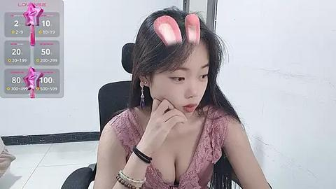 Media: Video of a young Asian woman with long black hair, wearing a pink lace dress, sitting in a black chair, in a white room with a temperature gauge, pink devil horns on her head.