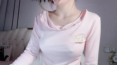 Media: Video of an Asian woman with fair skin, medium-sized breasts, wearing a pink long-sleeve top with a cartoon polar bear design, indoors with a beige sofa and a floral patterned blanket.