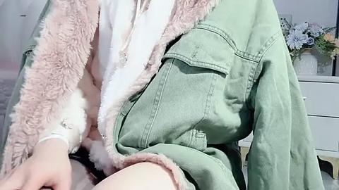 Media: Video of a person wearing a pale green jacket with a fluffy pink fur collar, white shirt, and a hint of light-colored pants. The background features a white dresser with a blue vase.