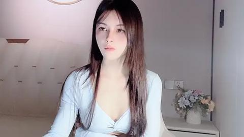 Media: Video of a young East Asian woman with long, straight brown hair, fair skin, and brown eyes, wearing a light blue V-neck shirt, seated in a modern, minimalist room with white walls and a flower arrangement on a table.