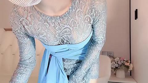 Media: Video of a woman wearing a sheer, lace, long-sleeved blue dress with a light blue belt. She is indoors, next to a white chair and a floral arrangement.