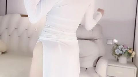 Media: A video of a slender woman in a sheer white dress, revealing her long legs and side, standing in a minimalist bedroom with a beige chair, white wall, and flower vase.