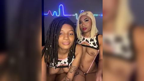 Media: Video of two women with medium skin tones, one with dreadlocks, the other with blonde hair. Both wear cow-patterned crop tops, revealing medium-sized breasts. Background features a neon heart light and blurred figures.