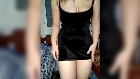 Media: Video of a woman in a black satin mini dress with thin straps, standing in a bedroom. The background shows a bed with a patterned duvet and a wooden headboard.