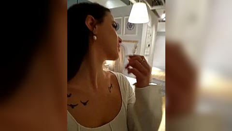 Media: Video of a woman with dark hair, wearing a white top, with a bird tattoo on her shoulder, standing in a modern, dimly lit room with white walls and a hanging lamp.