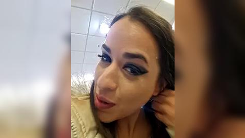 Media: Video of a light-skinned woman with long brown hair, wearing heavy black eyeliner and mascara, smiling in a selfie. She has a beige sweater and a blurred person in the background.